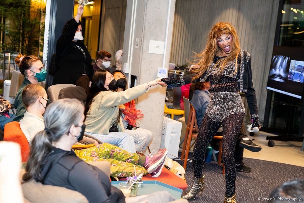  Iman, mid-performance, fully working the room. They've wandered into the crowd, and in this shot an audience member is reaching out, holding their hand, and they're mid-stride, their long hair flowing and moving like a rock star. They are head to toe sparkles and their stiletto ankle boots are sequined to the max.
