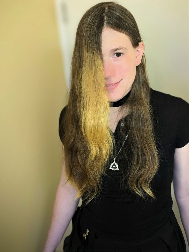 Ellie (me) in a black top, black skirt, therian necklace, and black choker. Her hair is in front of her face, brown with a blonde streak and tips!