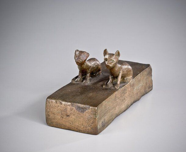 Animal coffin with the remains of two individuals. Bronze "brick" surmounted by two kittens seated next to each other, facing the long way, about halfway back. Facing them, the cat on the right is in a better state of preservation. His face is distinguishable. The cats have long, thin, almost exaggerated necks. Their tails lie on the block, curling around toward the left and ending by the front paws. The front legs are nearly vertical. The cats are resting on their derrieres. Their ears are in an "alert" position. Their bodies are incised with lines simulating fur, their toes with "toe" marks. The "brick" they are seated on is a tiny coffin for the cats.
