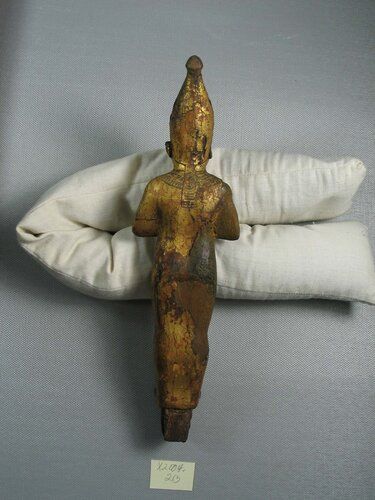 Standing mummiform figure of the God Osiris. Wood covered with gold leaf and having some bronze accessories.  The god wore the white crown flanked by feathers; the arms are indicated through the mummy wrapping and the hands exposed, knuckles meeting and each originally holding an attribute. The figure is provided with  a collar which meets behind, consists of two rows of vertically radiating beads, the lower ones are drop pendants. It is not apparent whether the vertical strips behind are a part of the collar or belong to the crown. They appear to hang from the crown.

Technic: The entire figure with white crown and a dowel on the under surface of the feet were carved out of a single block of wood. It is inserted in a small rectangular base with paneled sides which is of another single piece of wood. Near the four corners round holes were drilled from the bottom, reaching to within about 6 mm of the upper surface. Two of these holes were drilled too close to the sides and have broken through the panels. In the two front holes two of the round pegs remain which served to fasten the small base to its pedestal. The whole block was cut through to the bottom to receive the dowel in the feet of the figure and this shows below. 

The accessories separately added include 1) bronze uraeus on the front of the crown; 2) the feathers of the crown for which two dowel holes are visible on backside of each crown, 3) the eyes probably of glass or paste, 4) the beard and its straps (material not evident), 5) the attributes in the hands. The latter are closed and round holes are drilled for the reception of the fly-flap and scepter. The entire figure in each case received a layer of stucco on which gold leaf was laid. The details of the collar and ends behind modeled. The details of the collar were not carved in the wood but modeled in the plaster before the god leaf was applied.

Condition: The surface is considerably darkened (by resin?) thus the material of the eyes, and other accessories is difficult to determine. Some gold leaf and stucco has dropped off. The figure has lost feathers from the headdress, the beard and part of the inlay representing beard straps, both attributes, and its pedestal. This objecy has a worm-hole in the exposed wood of the right shoulder and the nose of is hopelessly flattened.The uraeus of 342 proves to be delicately engraved. The beard-straps were not metal but paste. The right eye looks like ivory to which the pupil was added in black paste (to a flattened, perhaps somewhat roughened surface); the left eye, however, is of glass. The cornea is whiter and the pupil blacker. Presumably the original left eye was lost that this is a repair.