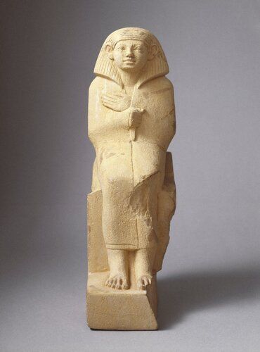 Yellow limestone statuette of a seated man. He wears a long cloak drawn tightly around body and grasped with right hand. Left hand extended flat on breast, palm down. Pleated wig. Simple block throne with plinth extending up to base of wig. Inscription on plinth, sides of throne and base. Name lost. 

Inscriptions: inscriptions on throne seem to be conventional offering formula texts. Inscription on plinth not entirely clear. “An offering which the king gives and Geb for” are the following words titles of the deceased?  
 
Condition: Broken at hips and assembled. Major portion of sides and back of throne missing. Portion of left foot missing. Area around left knee missing. Remains of paint, entire cloak originally painted pinkish-red. Wig and eyes painted black. 
