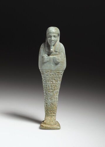 Pale green faience mummiform ushabti of Padineith. He wears a non-striated tripartite wig. His hands are crossed and juxtaposed on his chest, the right holding a mattock  and the left a pick. There is an incised bag behind left shoulder. There are eight lines of inscription on the front of the figure which continue with a single column of text on the back pillar.
Condition: The condition of the piece is good. The glaze is for the most part faded. The figure is wholly intact.