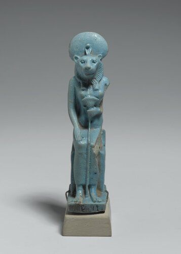 Pale blue-glazed faience figure of a lioness-headed goddess. She is seated upon a throne the sides of which are decorated with an overlapping scale-like feather pattern. Her right hand is upon her knees; her left hand holds a lotus to her chest. She is crowned with solar disk and uraeus.
Condition:  Left side of figure broken away.  Rear bears kiln rock marks. Chip in rear of sun disk.