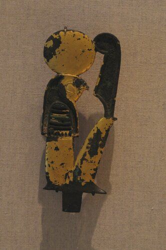 Gilt bronze relief figure of Horus (or Re-Harakhte?) mummyform with falcon head, seated and supporting large "m3't" feather on knees. Large sun-disk on head, tang on base and suspension loop on reverse of disk. Originally inlaid with glass. All details of reverse indicated by incisions. 

Condition: Extensive areas of gold-leaf missing. Only three pieces of original glass inlay preserved. A few scattered areas of corrosion.