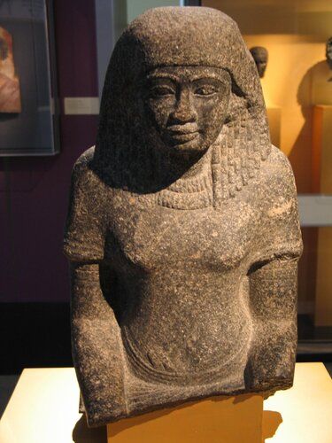 Dark grey granite bust of a seated man with striated and echeloned double wig, plastic eyebrows and plastic upper eyelid rims, double necklace, short sleeved over-garment, incised armlets, and on the right wrist a broad incised bracelet. The figure wears a pleated skirt with loop-fringed upped edge from which a double fold protrudes to the right of the navel. 
The back pillar bears two columns of inscription increasing to six columns on the back of the chair. On the front of the right shoulder there is an erased cartouche containing a name ending in “w’n-r” (Akhenaton or Amenhotep IV). A second erased cartouche on the right side of the chest is illegible.
Inscription on the rear of the seat and back pillar. The identity of this owner is as yet not established. Possibly Parennufer, Ramose, Kheruef, Nebamun, Amenemhat, Surer or Huy. 
Condition: Broken across wrists just below angle between garment and torso. Nose mostly missing, ears damaged; chipped and scratched in numerous places. Upper right and left rear corners of seat missing. Surface discolored; brownish black in spots.