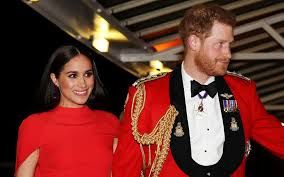 A photo of Harry & Meghan holding hands. Meghan is wearing a red caped evening dress and Harry is in his formal dress uniform.