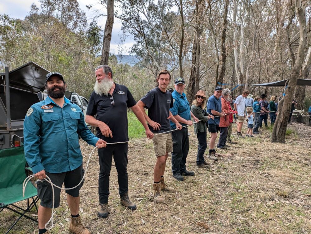 A line of people, holding a rope, on a riverbank 