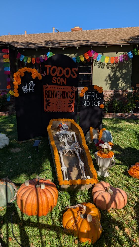 A beautiful display featuring pumpkins and marigolds with a skeleton in an open casket. The tombstones behind it have the LA finger sign made of skeleton hands, and text reading "todos son bienvenidos" right next to another stone reading "pero, no ICE."