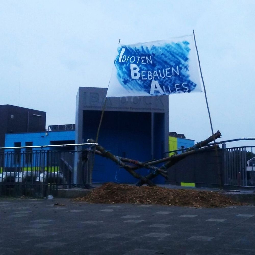 Foto vom Tor zum Übergang zum IBA Dock Bürogebäude. Große blaue Eingangstür, grau umrahmt. Im Hintergrund ist der bewölkte Himmel und das Bürogebäude zu sehen. Die Fassade des Gebäudes besteht aus blauen und grünen Kacheln. 
Vor dem Eingangstor liegen aufgehäufte Hackschnitzel. Der Zugang ist von Birkenstämmen blockiert. Über der Blockade weht ein Banner mit der weißen Aufschrift I dioten B ebauen A lles auf blauem Hintergrund.