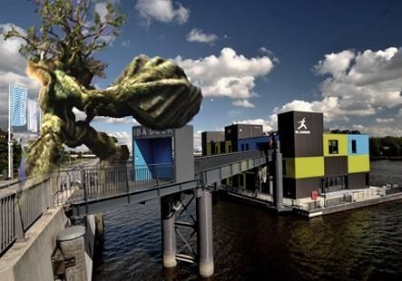 Ein Foto vom IBA Dock Bürogebäude. Das Gebäude steht auf einem schwimmenden Fundament. Unten ist das Wasser der Elbe zu sehen. Das Bild ist eine Seitenansicht. Links im Bild ist der Deich, mittig der Übergang und rechts das Bürogebäude zu sehen.  Links ins Bild wurde eine baumartiges Wesen mit einem Fotobearbeitungsprogramm hinzugefügt. Es handelt sich um ein Ent. Eine fiktionale Gestalt aus Herr der Ringe, mit einer Baumkrone als Kopf und baumstamm-artigem Körper. Das Ent ist sehr groß, es reicht über den oberen Bildrand hinaus. Es halt zudem einen großen Felsbrocken in den Händen. Damit sieht es aus also würde es das IBA Dock angreifen.