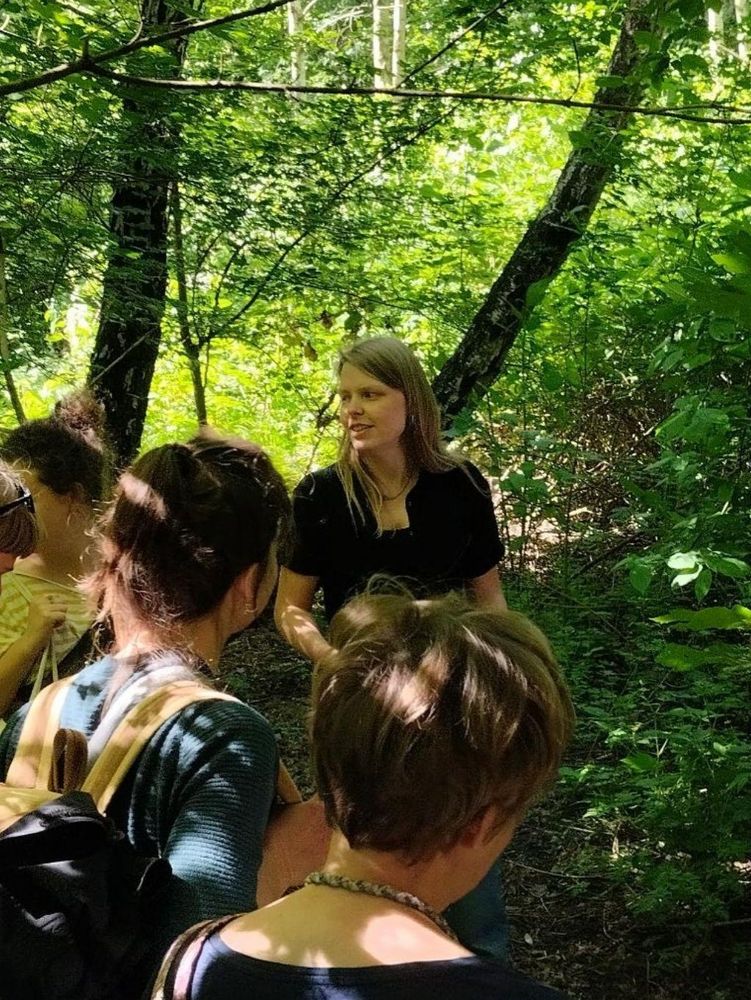 Vier Menschen hören einem Menschen zu, sie stehen im Wald, durch das dichte Laub scheint die Sonne.
