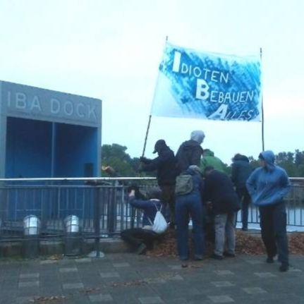 Ein Foto etwas seitlich vom Eingang zum IBA Dock. Etwa 8 Menschen sind vor der Eingangstür zu sehen. Sie befestigen Birkenstämme am Geländer. Bei ihren Füßen liegt ein Haufen Hackschnitzel. Ein Mensch ist zu sehen, wie er eine Bambusstange am Geländer befestigt. An der Bambusstange gespannt ist ein weißes Banner. Es ist blau angesprüht mit weißem Text. Darauf steht: I dioten B ebauen A lles.