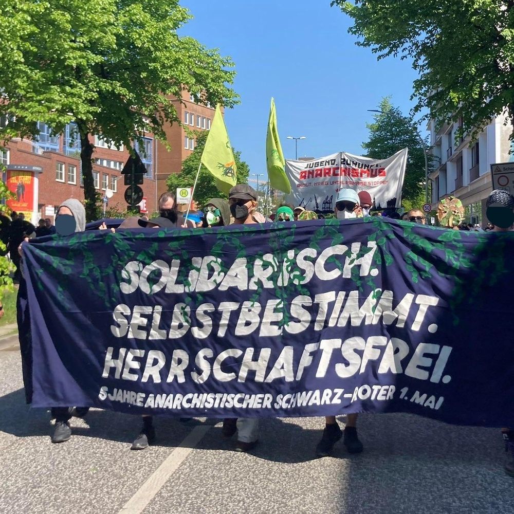 Fronttranspi mit der Aufschrift Solidarisch Selbstbestimmt Herrschaftsfrei. Dahinter Menschen mit grünen Blätter-Masken und WiWa-Bleibt Fahnen.