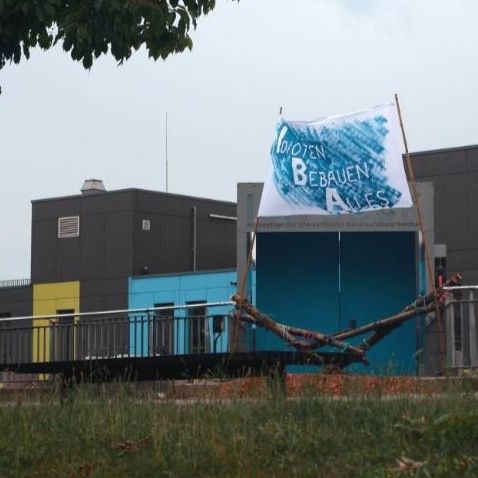 Ein Foto vom Eingang zum IBA Dock. Das Foto wurde von einer tiefergelegten Stelle aus gemacht. Im unteren Teil vom Bild ist etwas Wiese zu sehen. Im mittleren Teil ist die verbarrikadierte Eingangstür zu sehen. Es sind keine Menschen auf diesem Bild. Das blau besprühte Banner ist gut zu erkennen in der rechten Hälfte des Bildes. Im Hintergrund ist das IBA Bürogebäude zu sehen. Oben im Bild ist der bewölkte Himmel zu sehen.