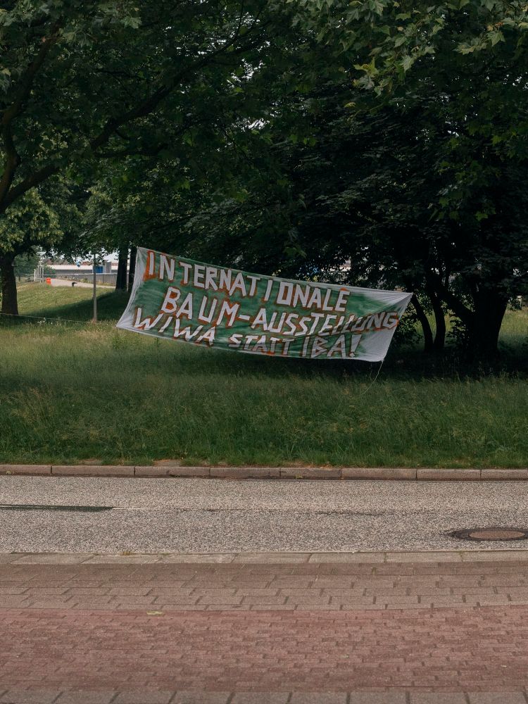 Foto zeigt ein Banner auf der gegenüberliegenden Seite einer Straße. Im Hintergrund ist eine Wiese und Gebüsch zu sehen. Das Banner ist zwischen 2 Bäumen gespannt. Das Banner grün und braun besprüht, die weiße Schrift sagt INTERNATIONALE BAUM-AUSSTELLUNG WIWA STATT IBA!