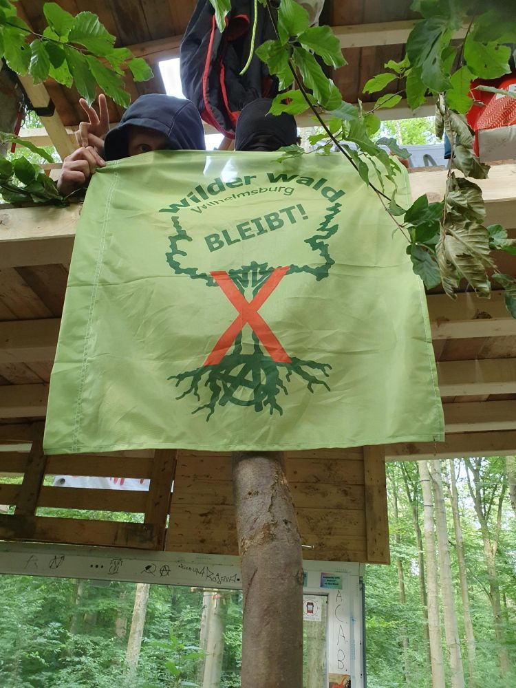Alt: Ein Quadratisches grünes Banner wird von zwei schwarz vermummten Menschen auf einer Holzplattform, die in einem Baum hängt gehalten. Darauf zu sehen ist das WiWaBleibt Logo mit dem orangenen X.