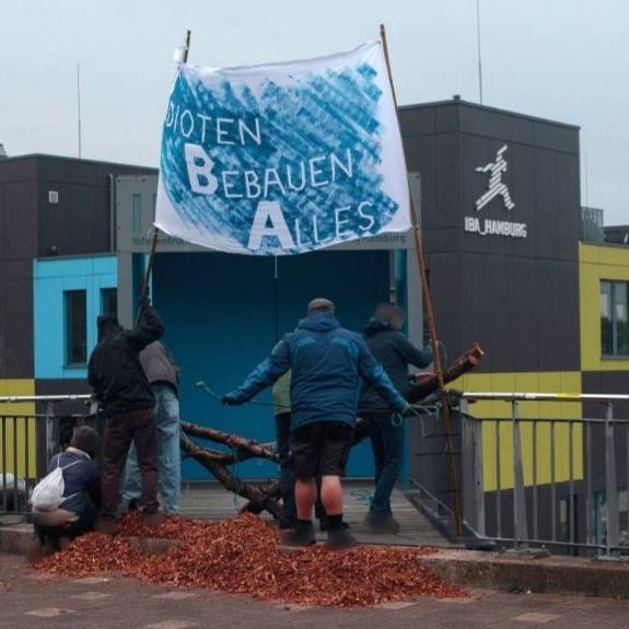 Ein Foto von vorne vom Eingang zum IBA Dock. Im Vordergrund sind 5 Menschen zu sehen. Einige befestigen links und rechts Birkenstämme. Damit blockieren sie den Weg zur Eingangstür. Zwei Menschen bringen Bambusstangen am Geländer an. Zwischen den Bambusstangen ist ein weißes Banner gespannt. Es ist blau angesprüht. Mit weißem Text steht draf: I dioten B ebauen A lles
Auf dem Boden im Vordergrund liegen Hackschnitzel aufgetürmt. Im Hintergrund ist das blau-grün gekachelte IBA Bürogebäude zu sehen.