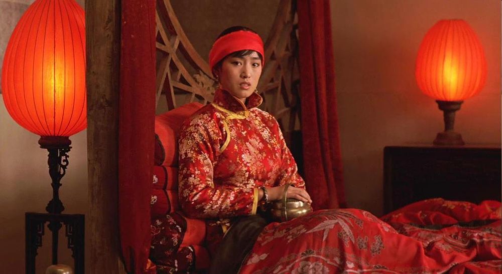 The inimitable Gong Li in a red collared robe and red headband sitting in a red bed holding a metal-looking hand warming thing and lamps with big red lampshades on either side. Gong Li is looking concerned, which she should, since her secret is known.