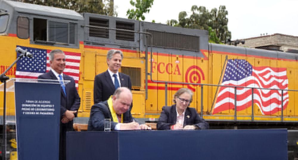 Two people (the mayor of Lima and the director of CalTrain) sitting at a table, signing an agreement, with two people (Peruvian transportation minister and US Secretary of State) standing behind them, with a diesel-electric locomotive covered in US flags in the background.