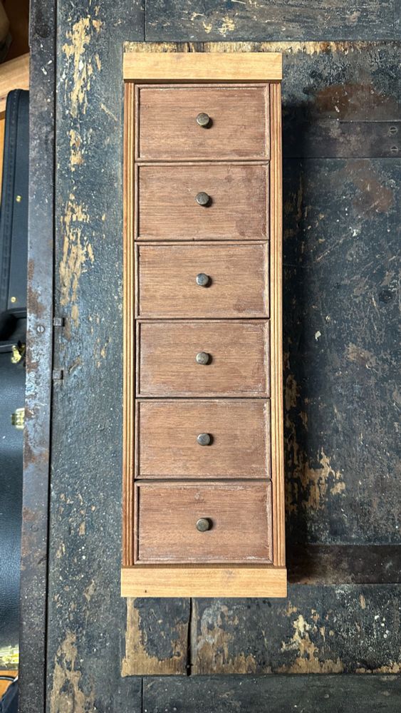 A tiny mahogany chest of drawers sitting on the vintage tool chest. It has six drawers about 3”x5” with cockbeading and a small brass knob on each drawer. The case sides are reeded. The molding is yet to be applied 