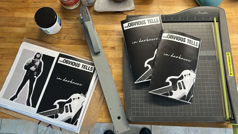 A long arm stapler next to a paper cutter, with to be assembled zines on the left and assembled on the right