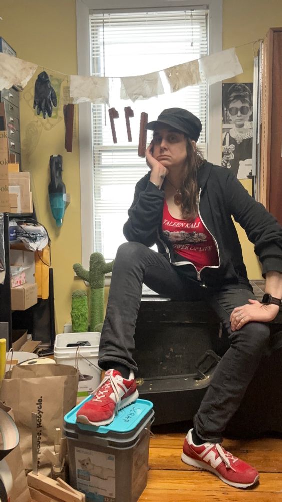 A greasy haired Jessica with her foot on the scrap metal bucket, surrounded by the detritus of the work room. A finishing line I behind her, with kerfing plane parts hanging letting their finish cure, and several rags with linseed oil on them drying enough they aren’t a danger of 
 combusting. They’re probably fine to throw away but why risk it. 