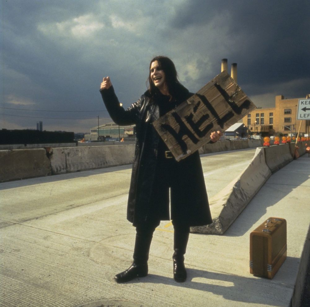 Ozzy hitchhiking with suitcase on a highway just outside of town, holding a sign that reads "HELL"