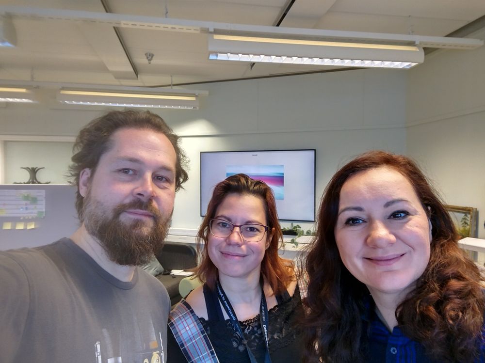 Three people looking straight at the camera, on the left a white man with beard,  middle and right white woman with red-ish longer hair.in the background a screen and office environment is visible 