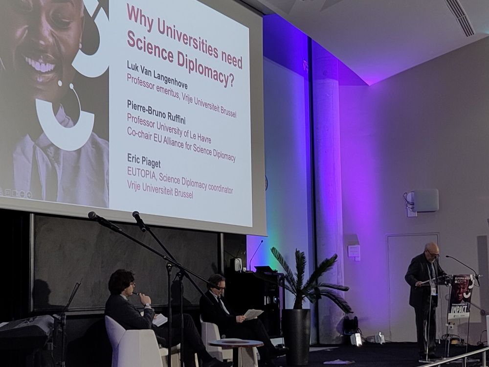 A stage showing two sitting white male researchers and one older white male academic talking. A slide in the background reads: why universities need science diplomacy and the names Luk van Langenhive, Pierre-Bruno Ruffini and Eric Piaget.
