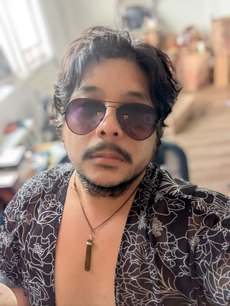 Jon Jon Selfie with a black shirt left open, flowery patterns in it on white. A cool pendant around their neck resting on her chest