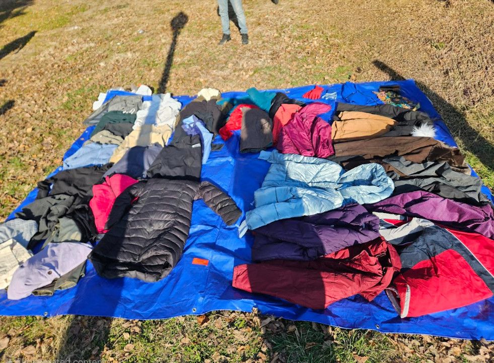 Collection of coats, jackets, and shirts sorted on a blue tarp outdoors.