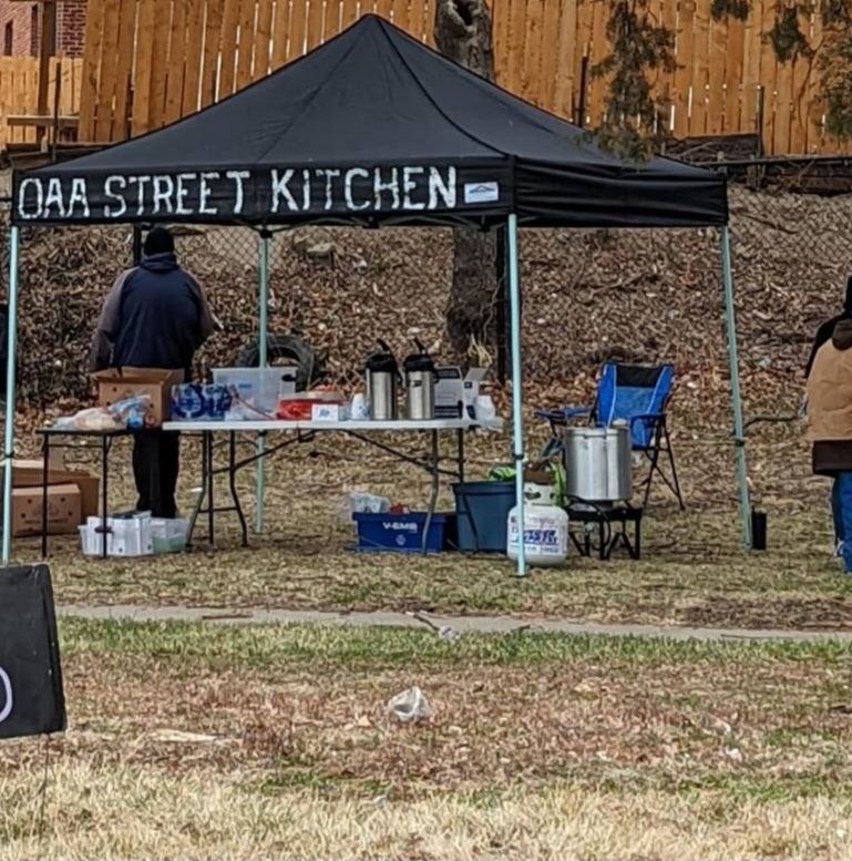 Tables with supplies next to a propane stove, all under a black tarp with words "OAA STREET KITCHEN" on it