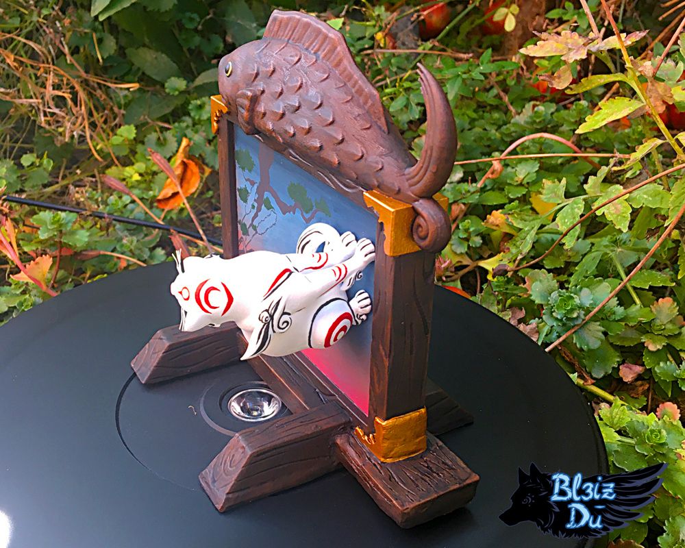 Photo of a figure set of Kabegami I made. Kabegami is a god from Okami, a white cat with red markings. He's sitting perpendicularly to a wooden board with a large painting in its centre and a wooden fish sculpture on top of the frame.