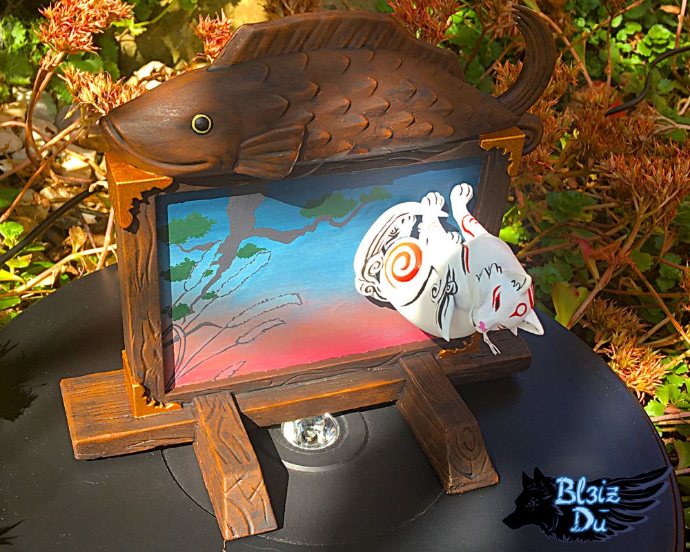 Photo of a figure set of Kabegami I made. Kabegami is a god from Okami, a white cat with red markings. He's sitting perpendicularly to a wooden board with a large painting in its centre and a wooden fish sculpture on top of the frame.