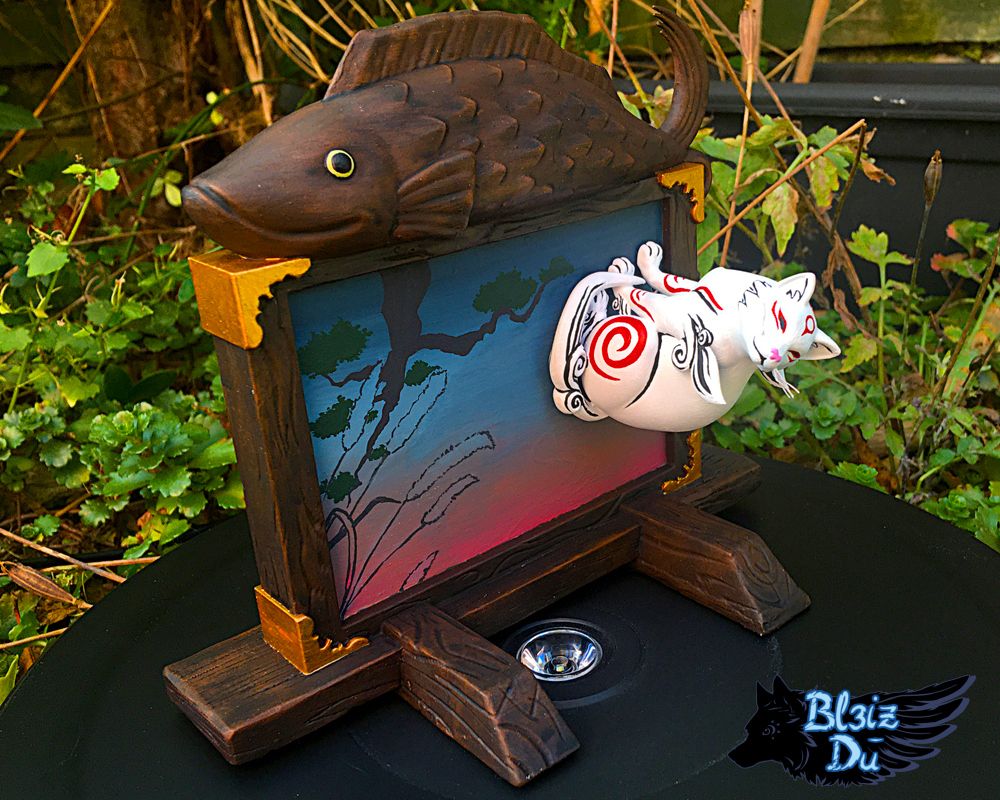 Photo of a figure set of Kabegami I made. Kabegami is a god from Okami, a white cat with red markings. He's sitting perpendicularly to a wooden board with a large painting in its centre and a wooden fish sculpture on top of the frame.