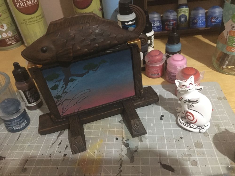 Photo of a figure set of Kabegami, the cat god from Okami. Kabegami is a white cat with red markings, sitting in front of a wooden board/wall with a wooden fish on top and a painting in the middle. Work in progress, still some painting left to do.