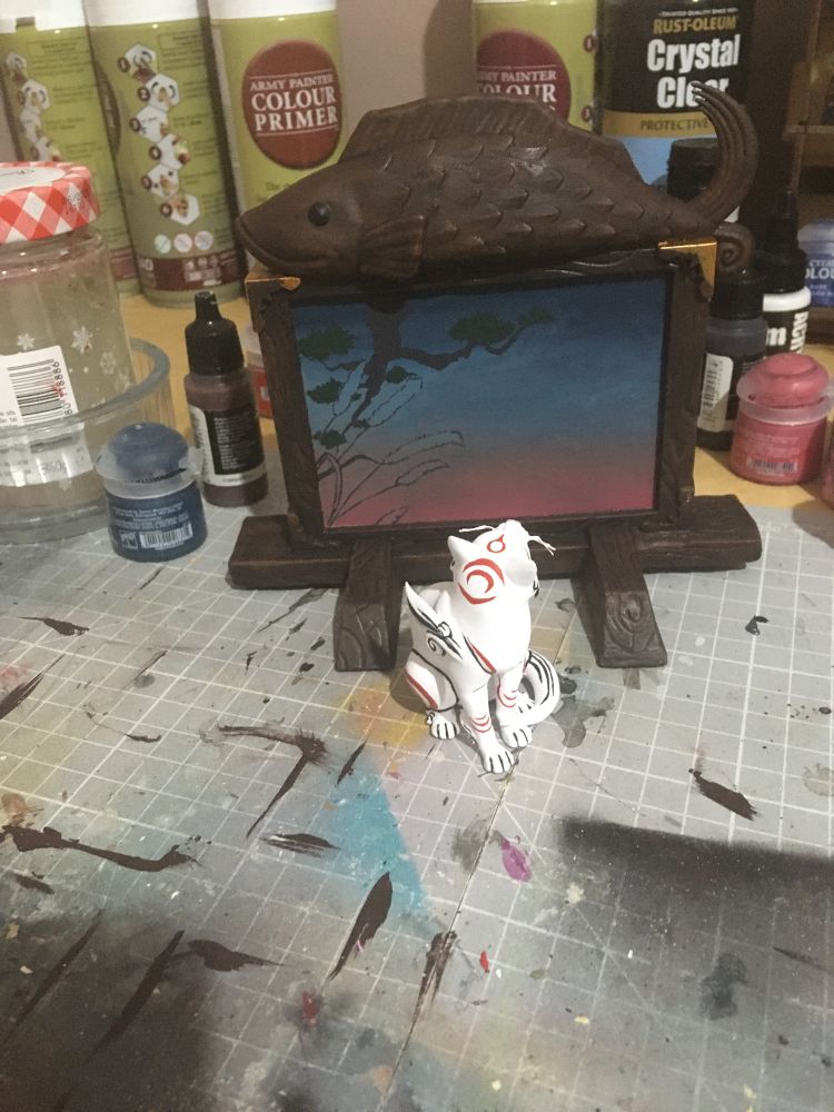 Photo of a figure set of Kabegami, the cat god from Okami. Kabegami is a white cat with red markings, sitting in front of a wooden board/wall with a wooden fish on top and a painting in the middle. Work in progress, still some painting left to do.