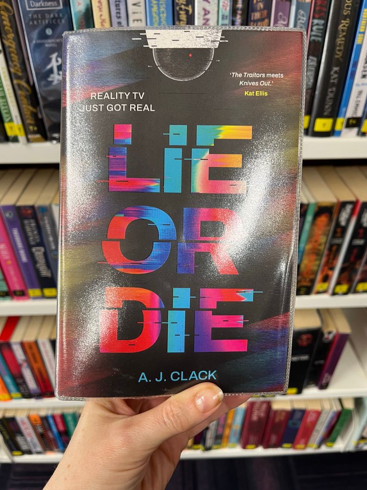 Cover of book ‘Lie or Die’ held up in front of library shelves 