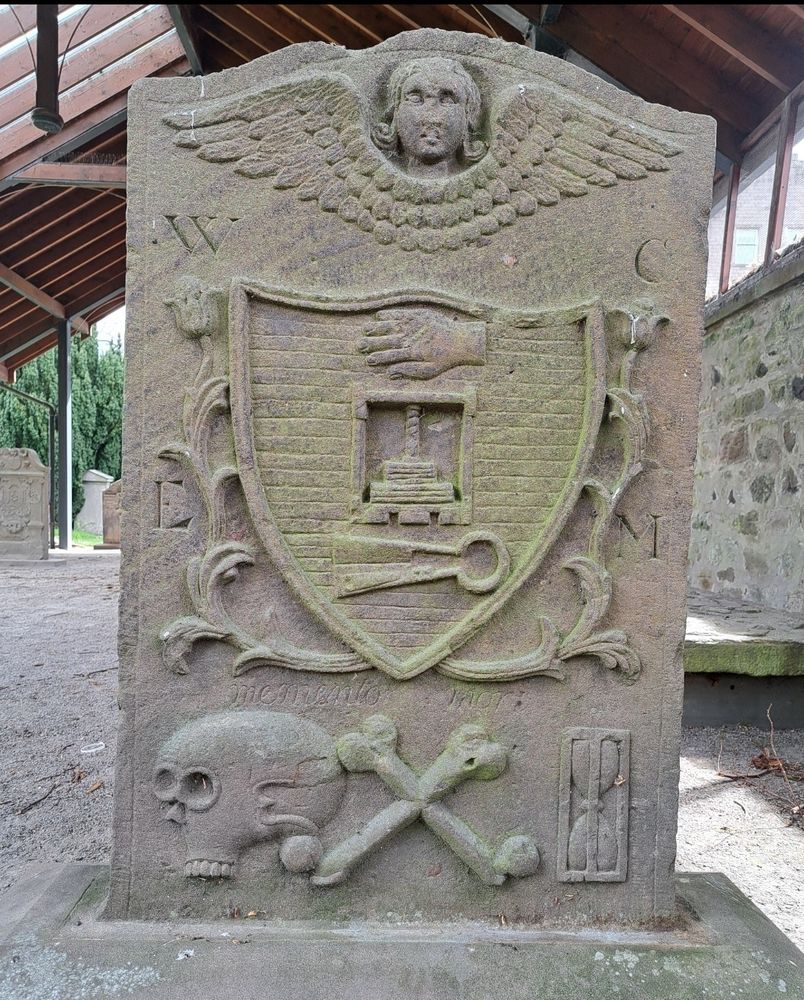 Photograph of the the 1788 gravestone of William Clement - a Dyer - in Greyfriars Burial Ground, Perth, Scotland.
A hand & a shears appear in the middle. An angel head & wings on the top. Memento mori symbols - a skull, crossbones, an hourglass - on the bottom.
https://www.undiscoveredscotland.co.uk/perth/greyfriars/index.html