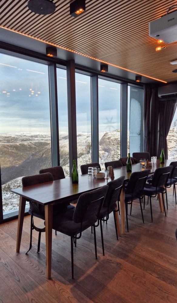 The image shows a modern dining or meeting room with a wooden table and chairs. The room has large floor-to-ceiling windows offering a panoramic view of a snowy mountainous landscape. The ceiling features a slatted wood design with embedded warm lighting.