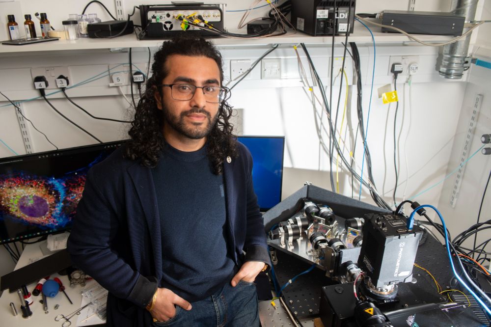 Akaash Kumar stands next to his multispectral imaging hardware which features several cameras. Behind him, a computer screen shows a colourful cellular image captured with his new microscopy system.