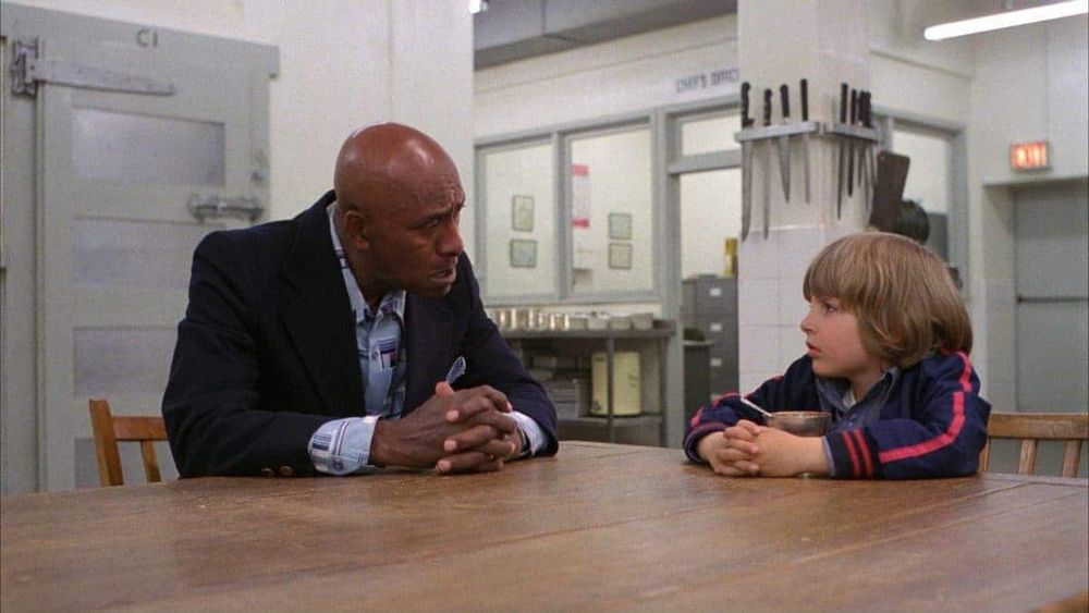 Dick Halloran and Danny Torrance speaking in the Overlook Hotel kitchen, with an array of knives hanging behind Danny, pointed toward his head.