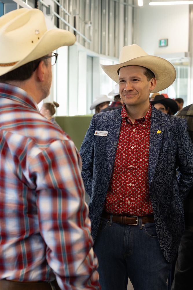 A picture of two men wearing cowboy hats. The person in the foreground is wearing a red and white plaid shirt. The man on the right is wearing a blue paisley blazer and red patterned shirt.