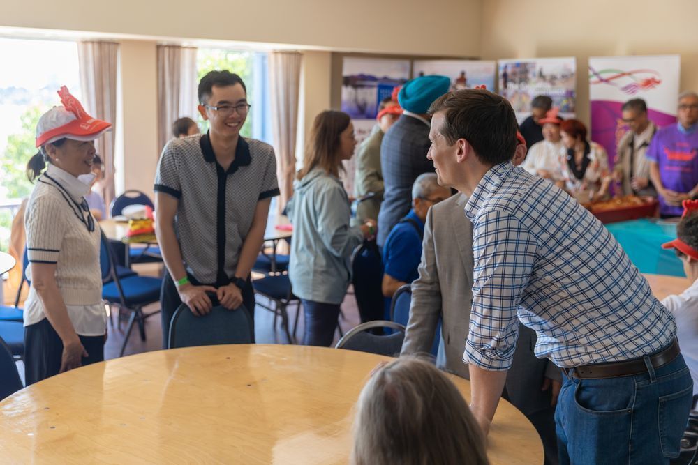 Jeromy Farkas leans forward on a table while smiling and speaking with attendees at a community event. People are gathered around the room chatting, with colourful banners and natural light in the background. 