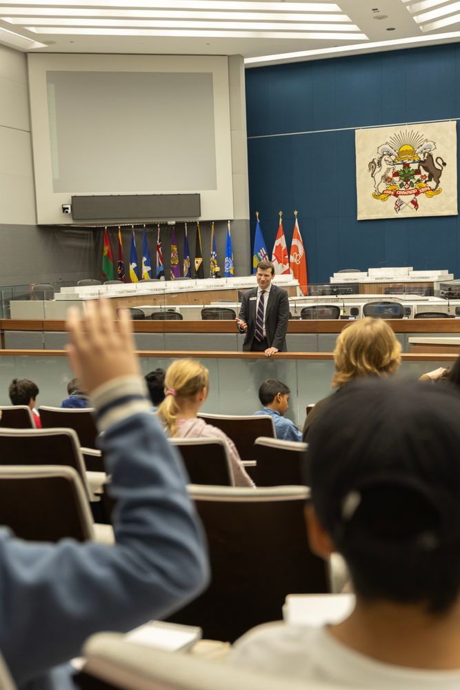 Picture of a man standing in front of a group of children seated. He is wearing a grey suit with a tie. Behind him are is a white and blue wall with a crest and several flags.