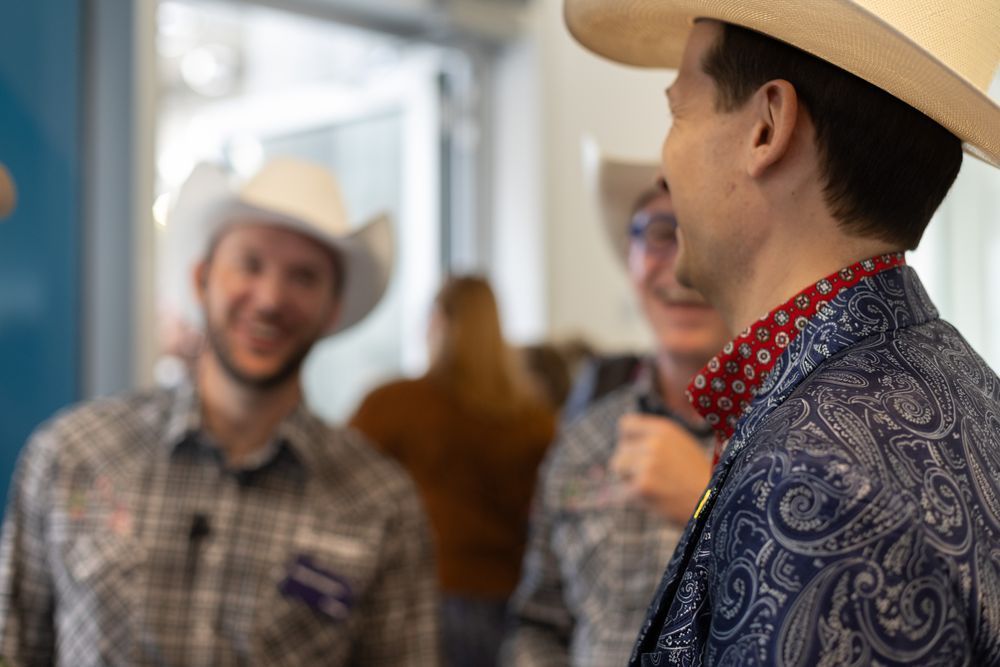 A picture of three men wearing cowboy hats laughing. The man on the left and middle are blurred and the man on the right is in focus, wearing a blue paisley blazer and red patterned shirt.