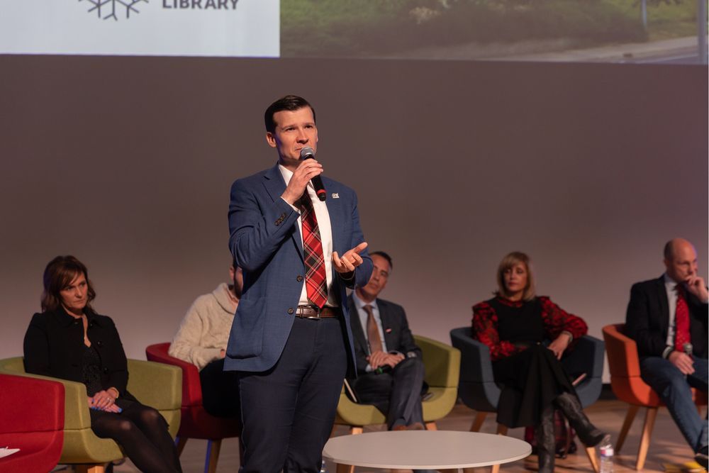 Picture of a man with a blue blazer, red turban tie, white dress shirt and navy blue pants talking into a microphone while five people sit behind him