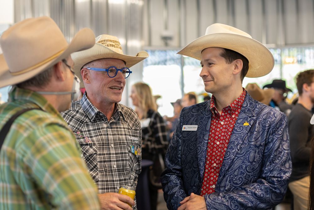 Picture of three men wearing cowboy hats. The man on the right is wearing a green plaid shirt and a respirator mask, the man in the middle is wearing a black and white plaid shirt and blue glasses and the man on the right is wearing a blue paisley blazer and a red patterned shirt.