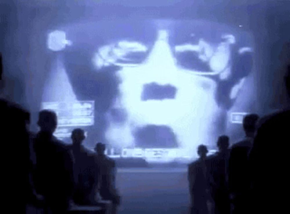 a group of people are standing in front of a large screen with a picture of a man 's face projected on it .