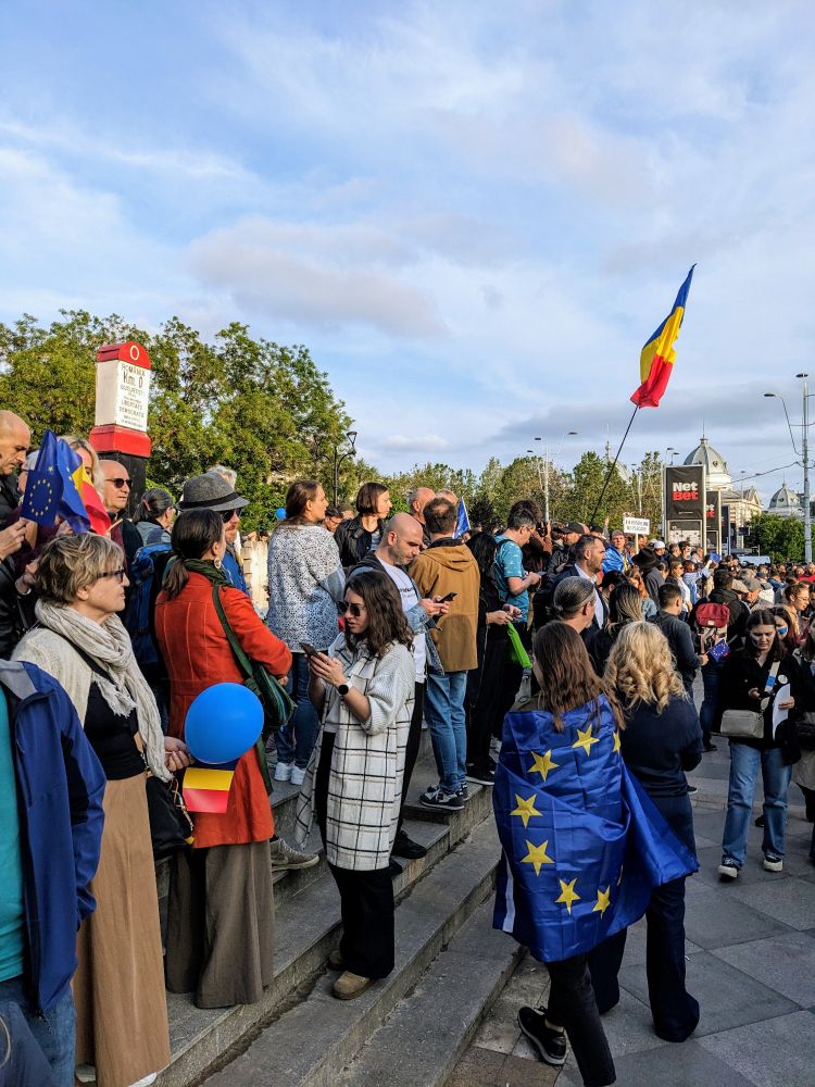 People gathered with blue balloons, Romanian flags, and someone wearing the EU flag 